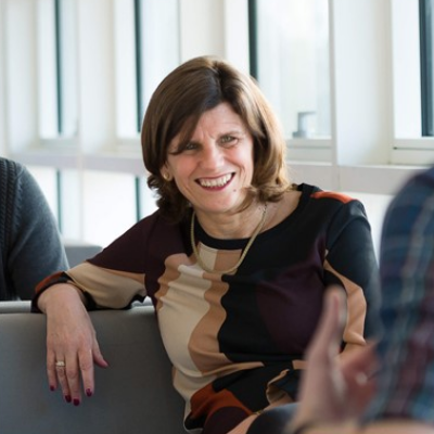 Person smiling while sitting in a bright room with large windows.