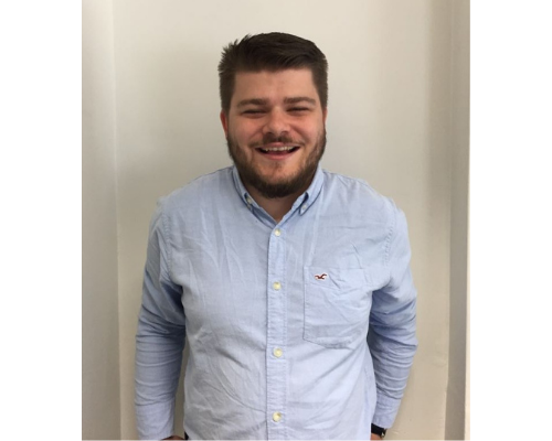 Person smiling, wearing a light blue shirt standing against a white wall.