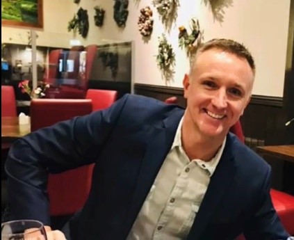 Smiling person in a restaurant, sitting at a table with red chairs and wall decorations in the background.