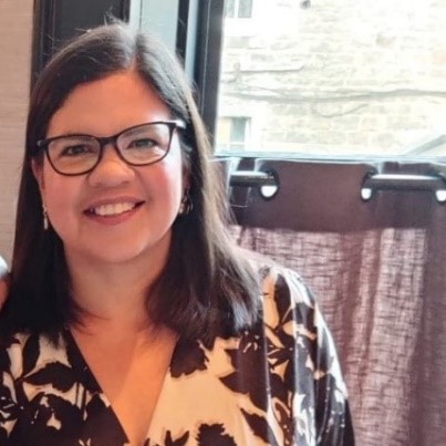 Person smiling indoors wearing glasses and a patterned shirt.
