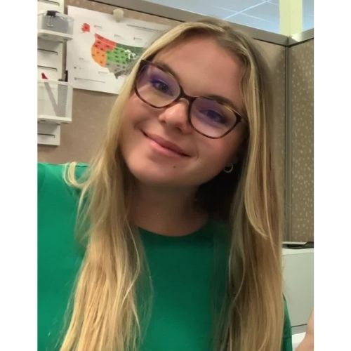 Person smiling in an office cubicle wearing glasses and a green shirt.