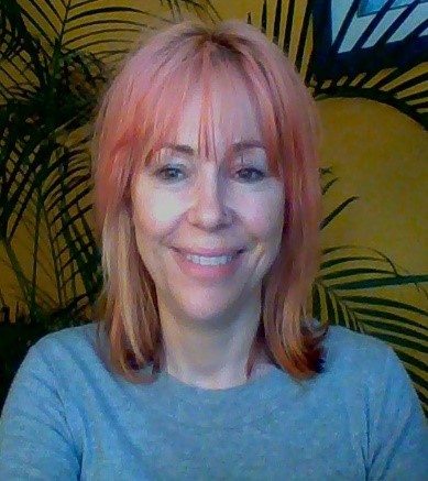 Person with pink hair smiling in front of plants and a yellow wall.