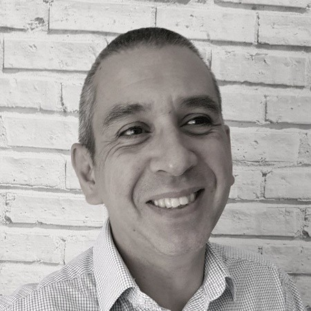 Person smiling in front of a white brick wall, wearing a checkered shirt.