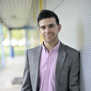Person in a suit smiling outdoors with a blurred background.