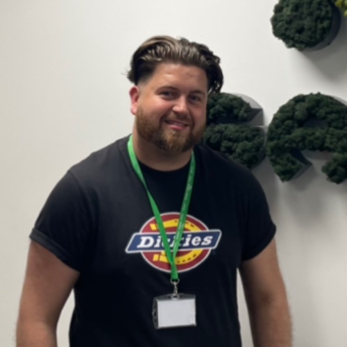 Smiling individual wearing a black t-shirt with logo and a green lanyard, standing indoors with greenery in the background.
