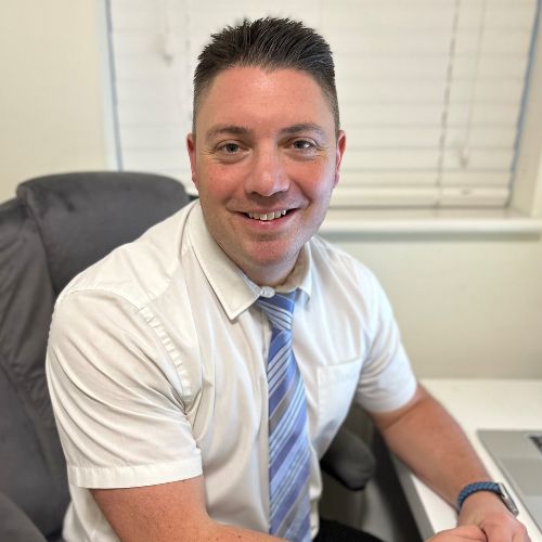 Person smiling while seated at a desk, wearing a white shirt and blue tie.