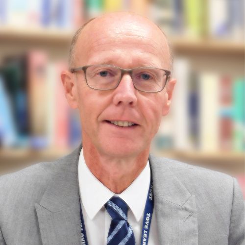 Person in glasses wearing a suit and tie, standing in a library.