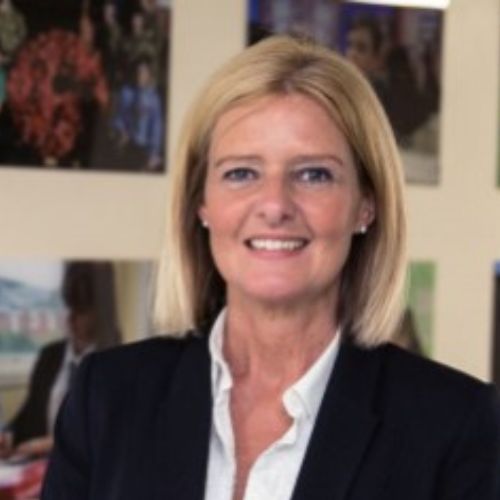 A person with light hair smiling in an office setting with photos on the wall.