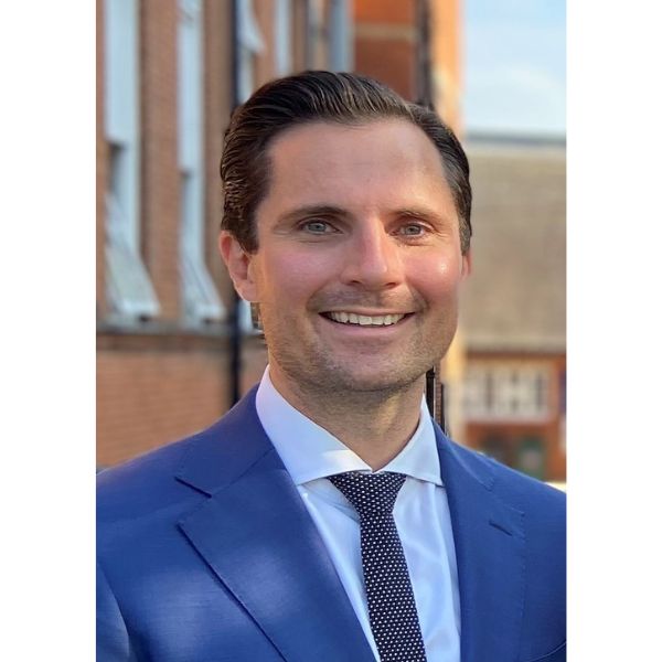 Person in a blue suit and polka dot tie smiles outdoors.