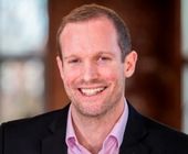 Person smiling in a blurred indoor setting, wearing a suit and pink shirt.