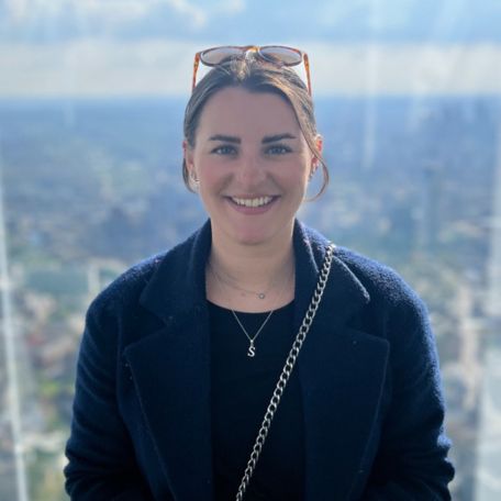 A person smiling with a cityscape in the background on a sunny day.