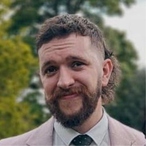 Person smiling with a trimmed beard, wearing a light suit outdoors.