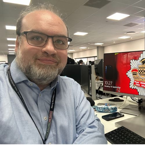 Person smiling at a desk with computer monitors in a busy office.