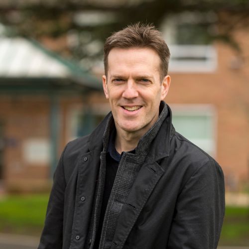 Person smiling outdoors in a black jacket, with blurred building in the background.