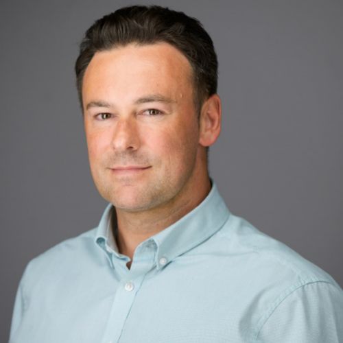 Person with short dark hair in a light blue shirt, smiling slightly against a grey background.
