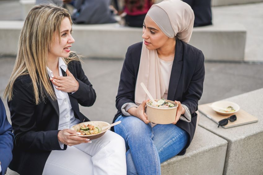 Two young women sitting outdoors on a stone bench, having a conversation over lunch