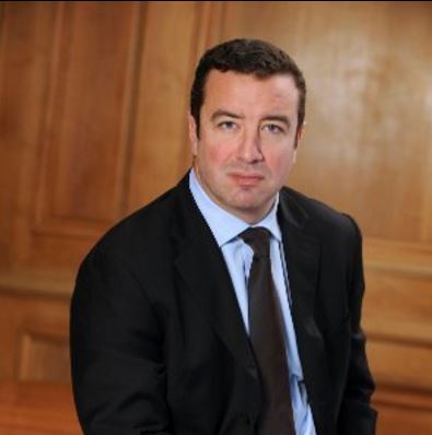 Person in a suit with a wooden panel background.