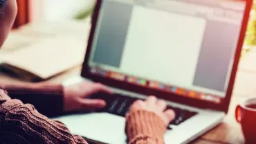 Person typing on a laptop at a wooden desk with a red mug