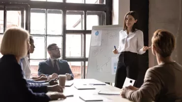 Person presenting to a group in a conference room with a whiteboard and notes.