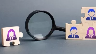 Wooden puzzle pieces with people icons next to a magnifying glass.