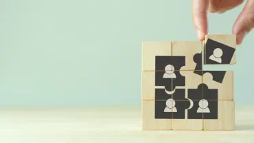 Hand placing a puzzle piece on wooden blocks with people icons.