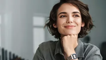 Person with short hair smiling, wearing a wristwatch, thoughtful pose.