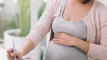 A person holding their pregnant belly while writing at a desk.