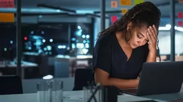 Person sitting at a desk with a laptop, appearing thoughtful in a dimly lit office.