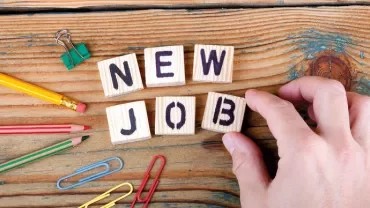 Wooden blocks spell 'New Job' on a wooden surface with office supplies nearby.