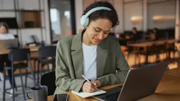 Person wearing headphones writing in a notebook with a laptop in a shared workspace.