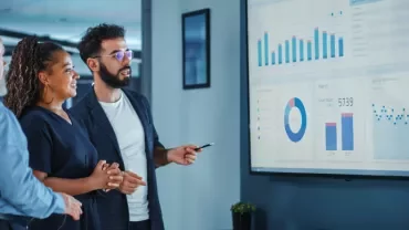 Three people discussing statistics on a large screen in an office.