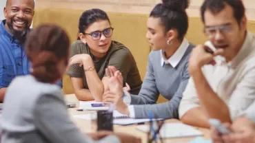Group of people having a discussion at a table, with notebooks and devices.