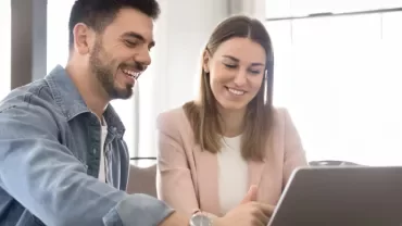 Two people are sitting at a table facing a laptop while smiling. 