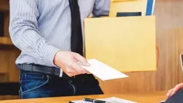 An employee, wearing a shirt and tie, extends to their boss while holding a box of documents. 