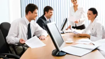 Group of people in a meeting around a conference table with documents and a computer.