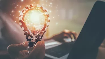Person holding a glowing lightbulb with a brain, symbolizing creativity, next to a laptop.