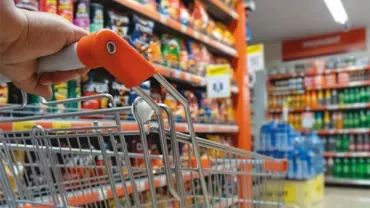 A hand pushing a shopping cart in a grocery store aisle filled with colorful products.