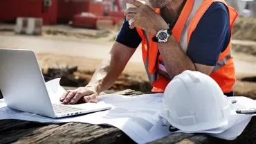 Person in a construction site reviewing blueprints and using a laptop.