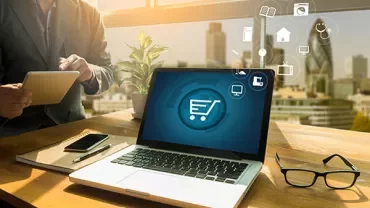 Person with laptop displaying shopping cart icon, using tablet, glasses nearby.