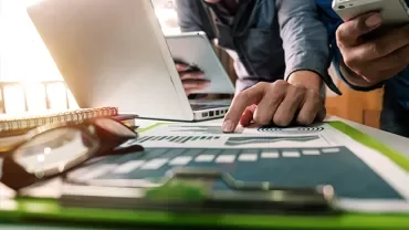 Two individuals collaborating with a laptop, smartphone, and documents on a desk.