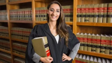 Person in academic gown holding a book in a library.