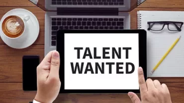 Tablet displaying 'Talent Wanted' on a desk with laptop, coffee, notepad, and glasses.