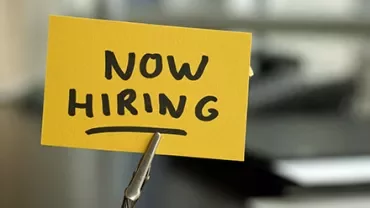 Sign with 'Now Hiring' in bold letters clipped on a desk.