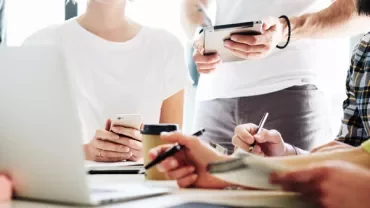 People collaborating at a table with laptops, phones, and notepads.