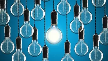Numerous hanging light bulbs with one lit against a blue background.