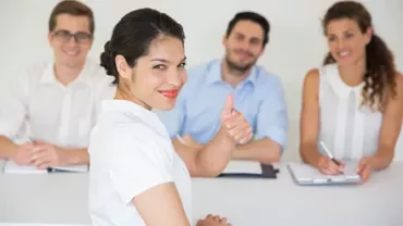 Person giving a thumbs up at a meeting with three people smiling in the background.
