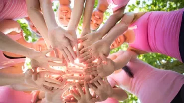 Group of people in pink tops with hands joined in a circle, sunlight shining through.