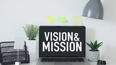 Laptop on desk displaying 'Vision & Mission' text.