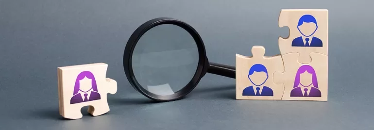 Wooden puzzle pieces with people icons next to a magnifying glass.
