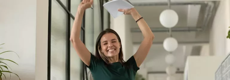 Person celebrating in an office, holding a notebook with arms raised.